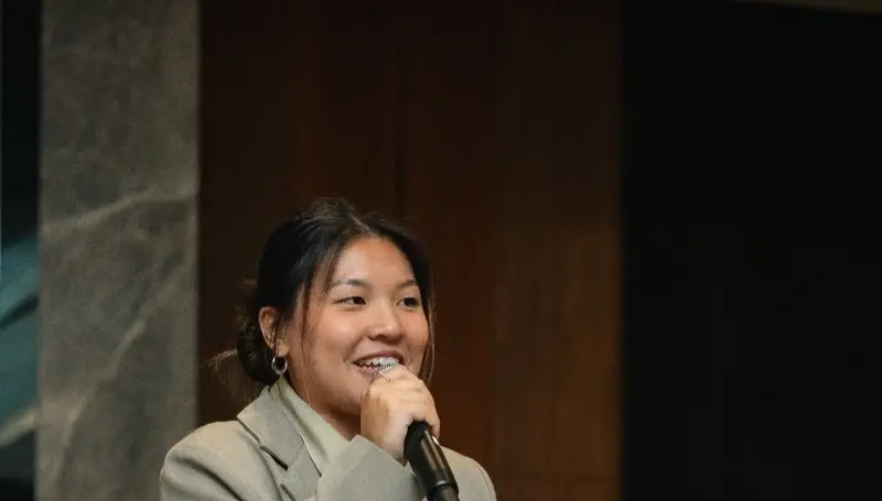 A female emcee in a grey blazer smiling as she speaks into a microphone at a hotel function