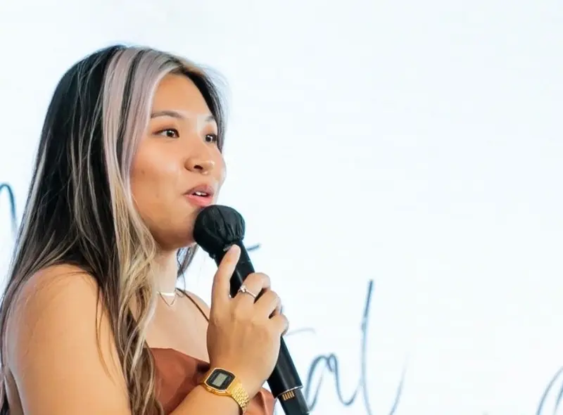 A female host in a satin dress speaking into a microphone in front of a white projection screen