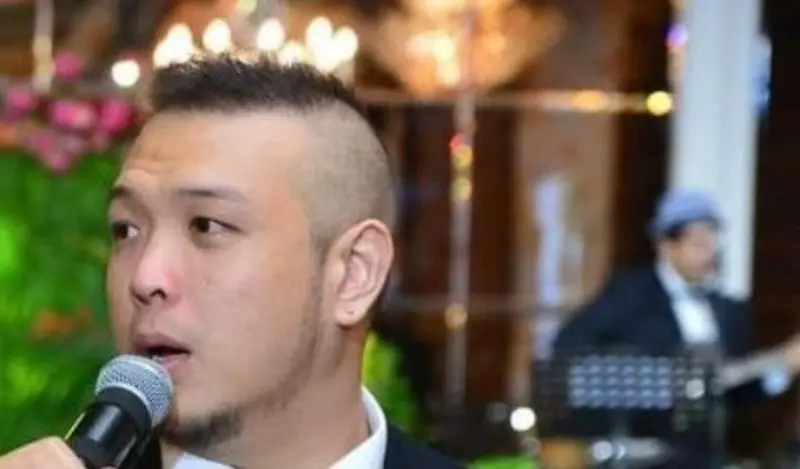 A male emcee in a tuxedo and bow tie speaking into a microphone at a wedding reception with a flower wall behind